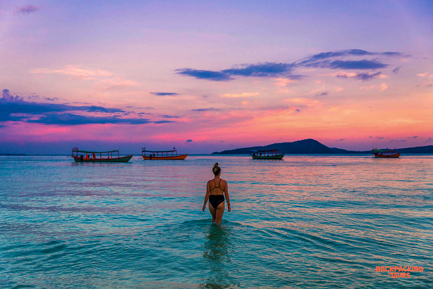 A woman wades into the calm, colorful waters of Koh Rong at a vibrant sunset.