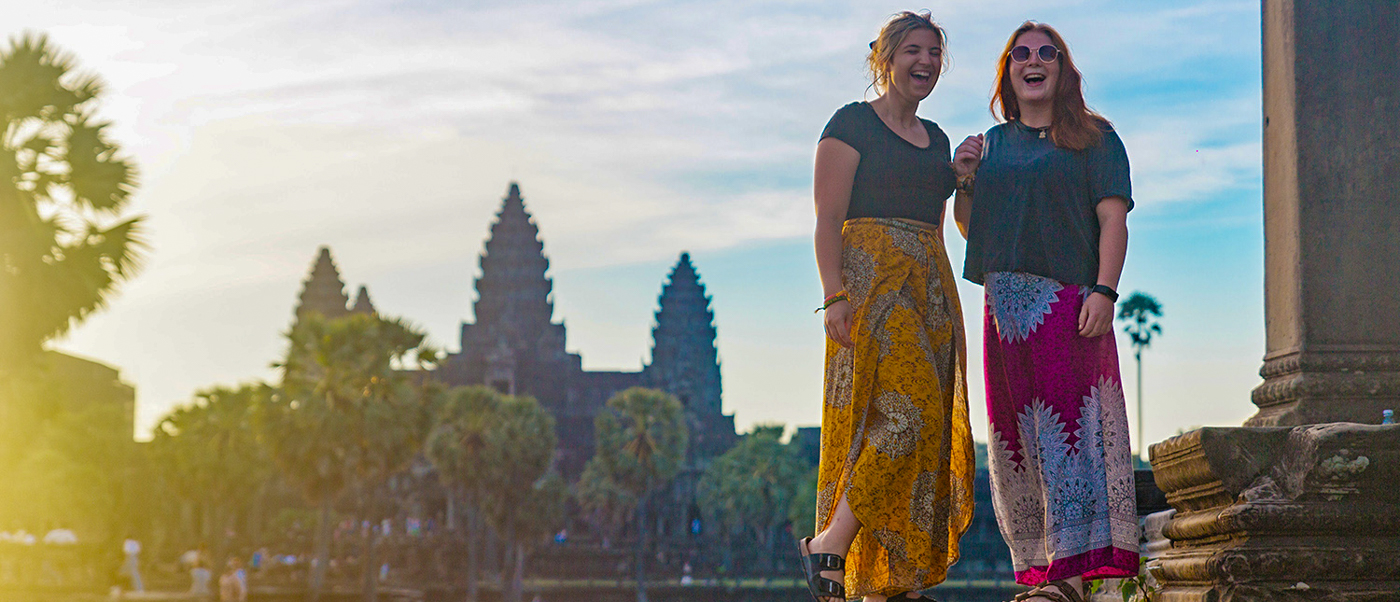 Two happy women in patterned skirts laughing with the iconic Angkor Wat temple in the background.