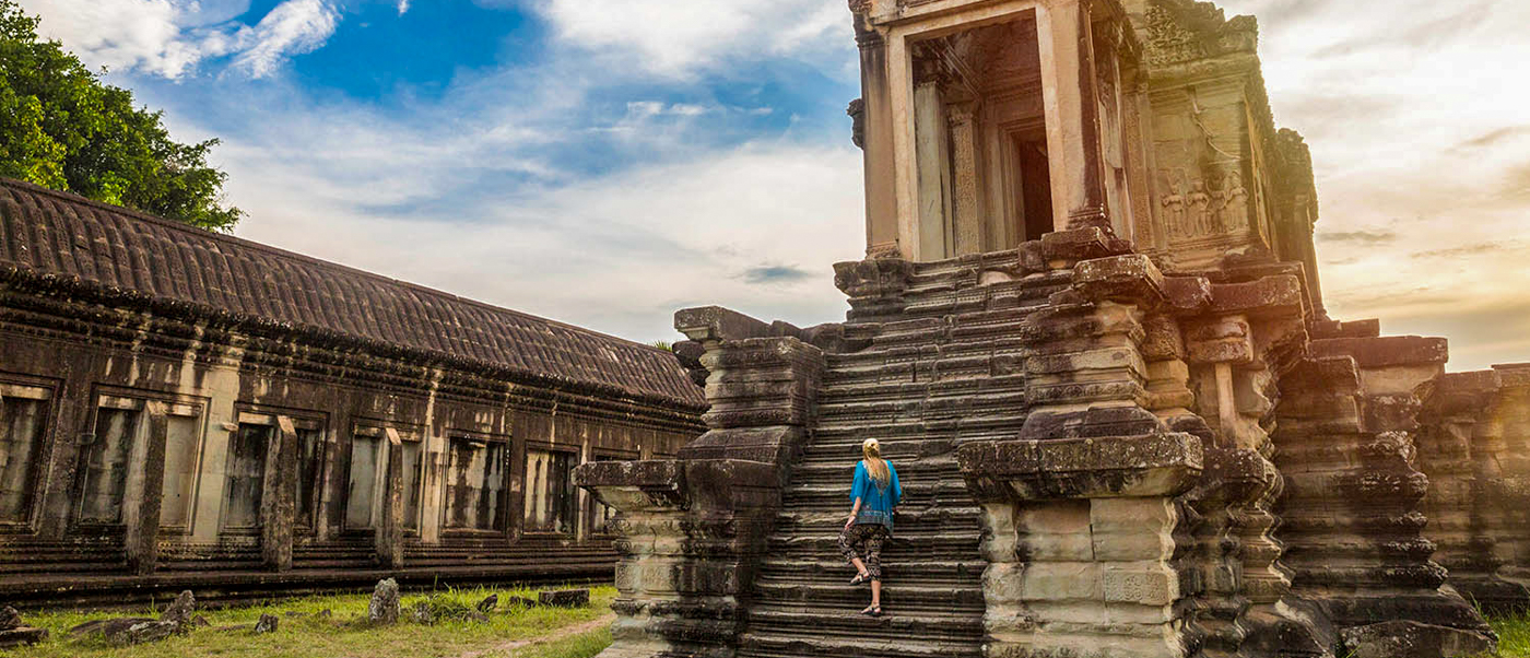 A traveler climbs the steep, ancient stone steps of Angkor Wat temple under a bright sky.
