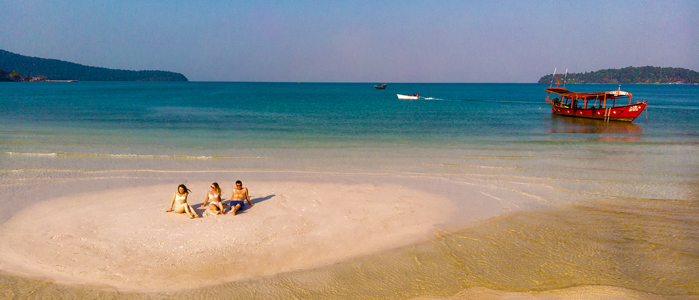 Three friends relax on a sandy island beach in Koh Rong with a red boat nearby.
