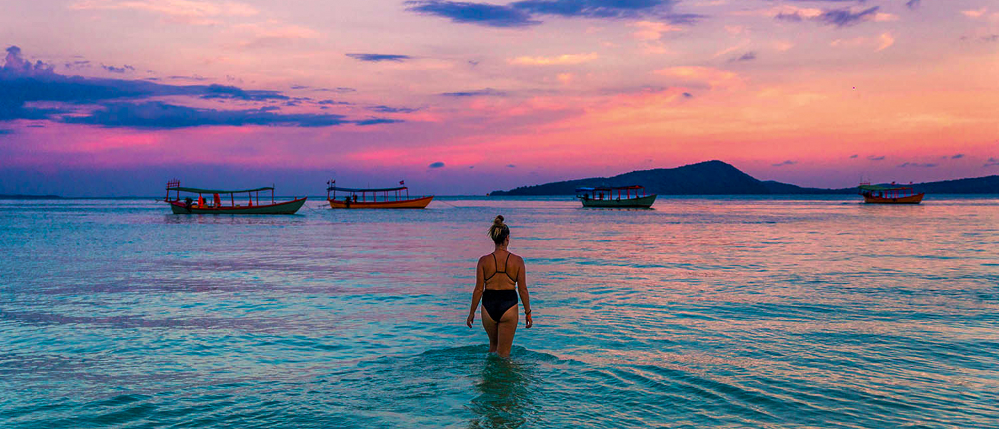 A woman wades into the clear turquoise water of Koh Rong during a vibrant sunset.