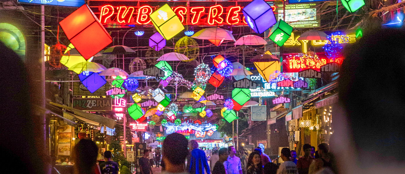 Lively Pub Street in Siem Reap at night, with colorful lanterns, umbrellas, and neon signs illuminating people.