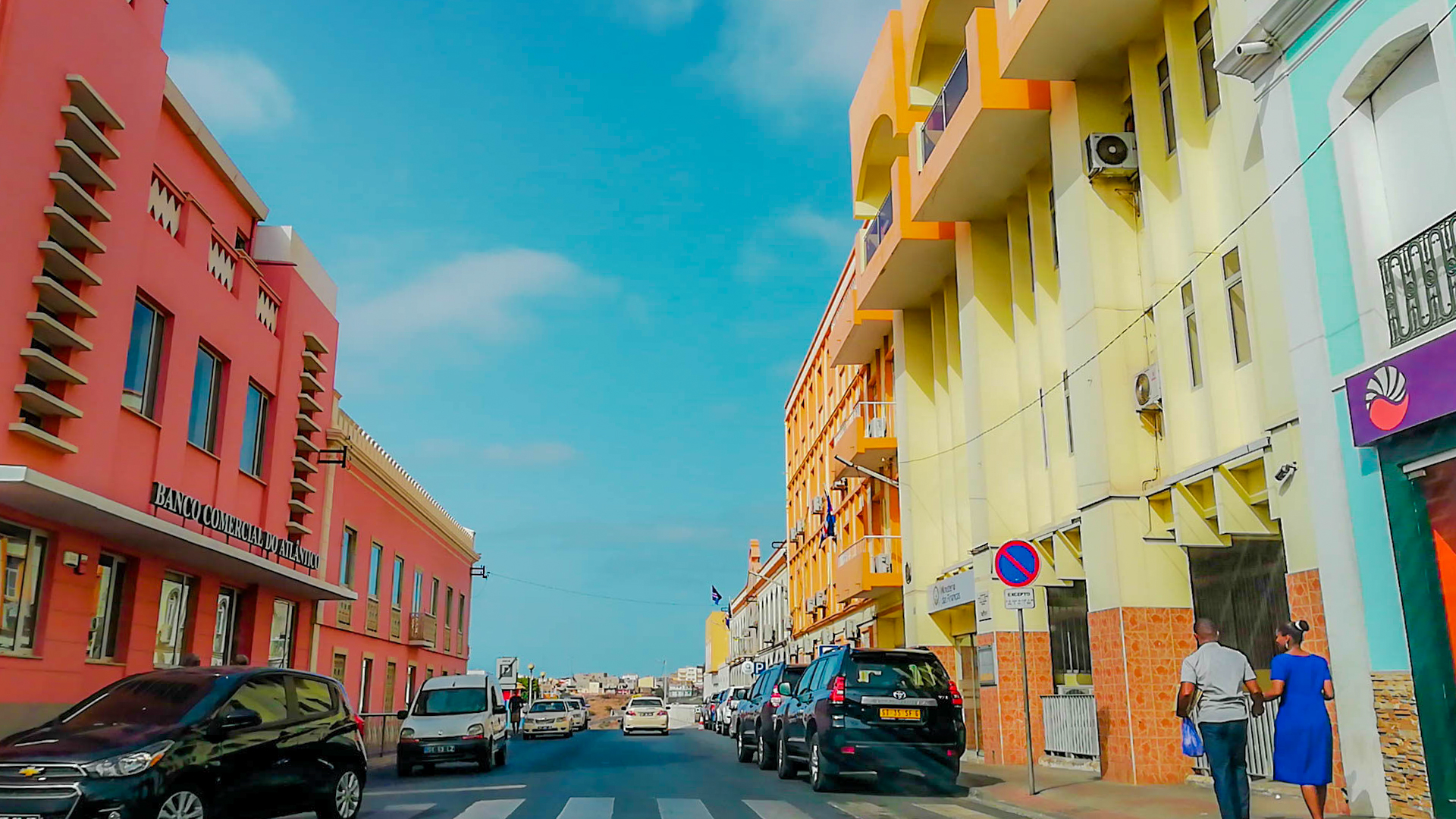 Vibrant pink and yellow buildings line a busy street with cars and pedestrians in Cape Verde.