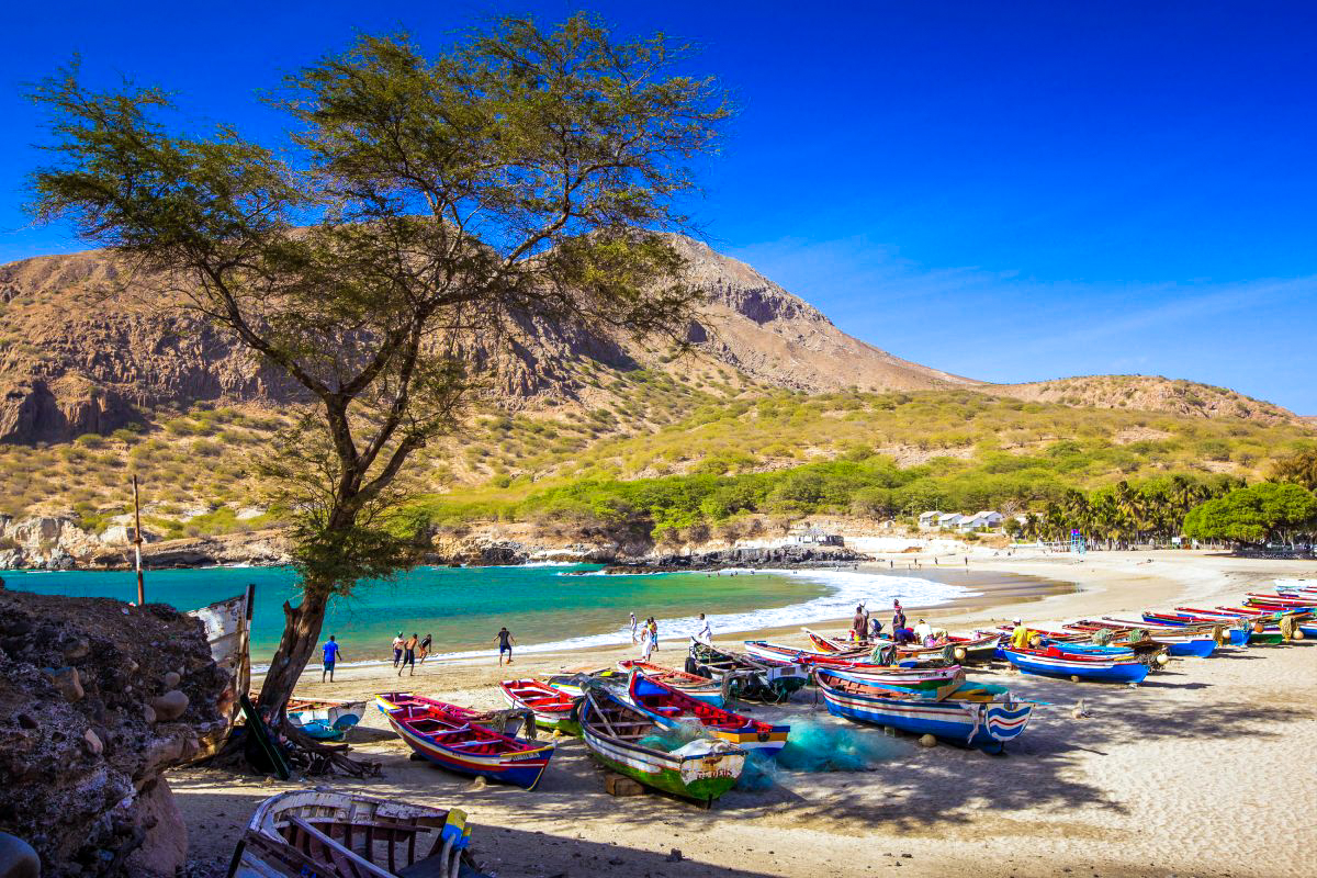 Colorful fishing boats line a sunny beach with turquoise water and volcanic mountains in Cape Verde.