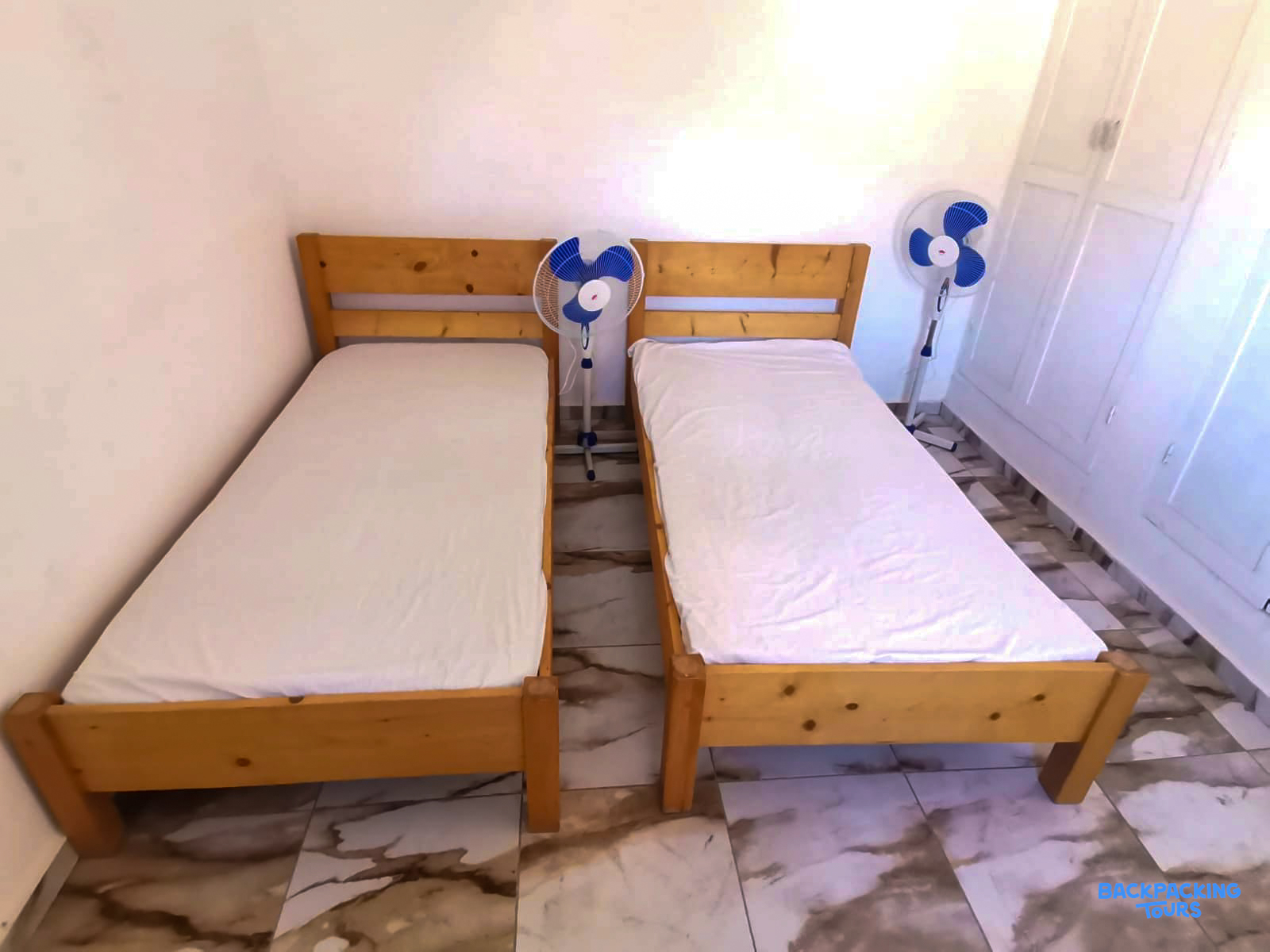 A bright, clean room featuring two single wooden beds with white bedding and standing fans.