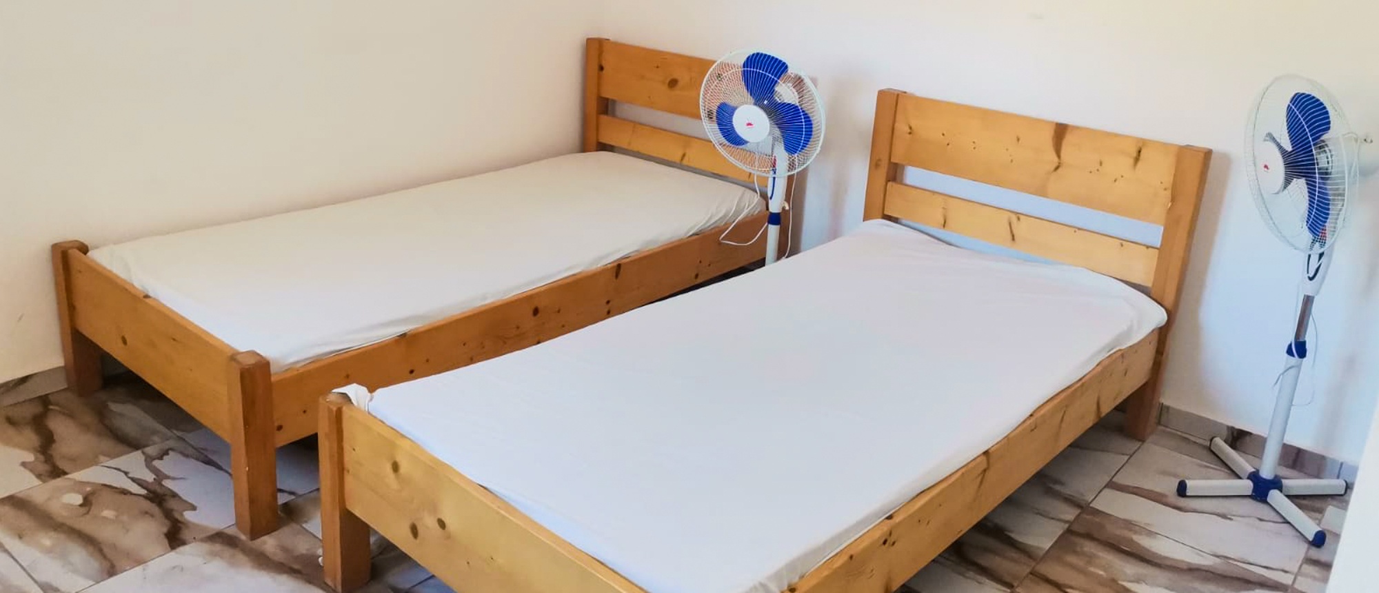A simple bedroom with two single wooden beds, white sheets, and two standing fans.