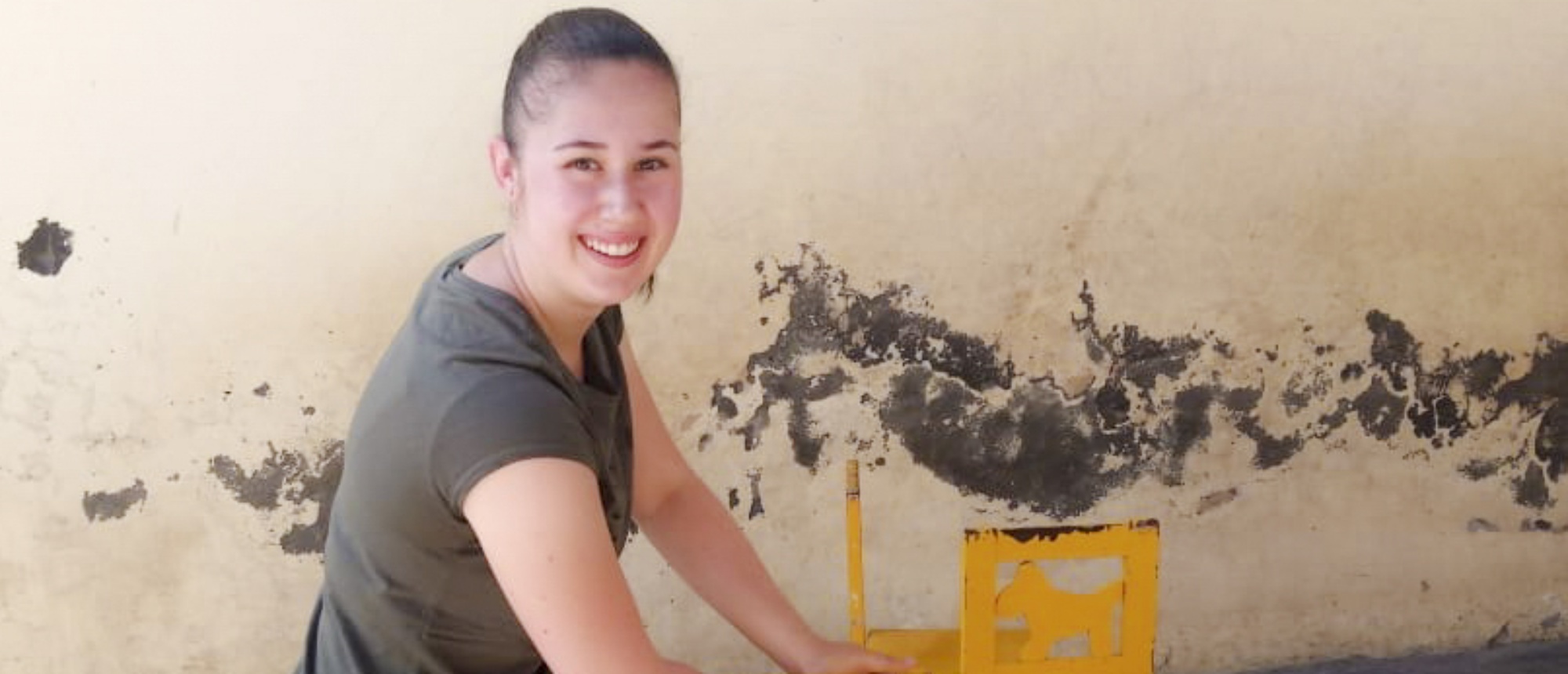 A smiling woman in a dark shirt working on a damaged wall with a yellow object.