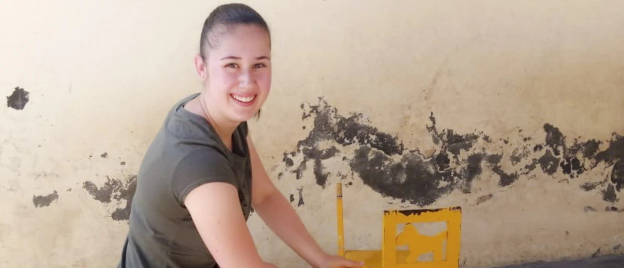 A smiling woman in a dark shirt working on a damaged wall with a yellow object.