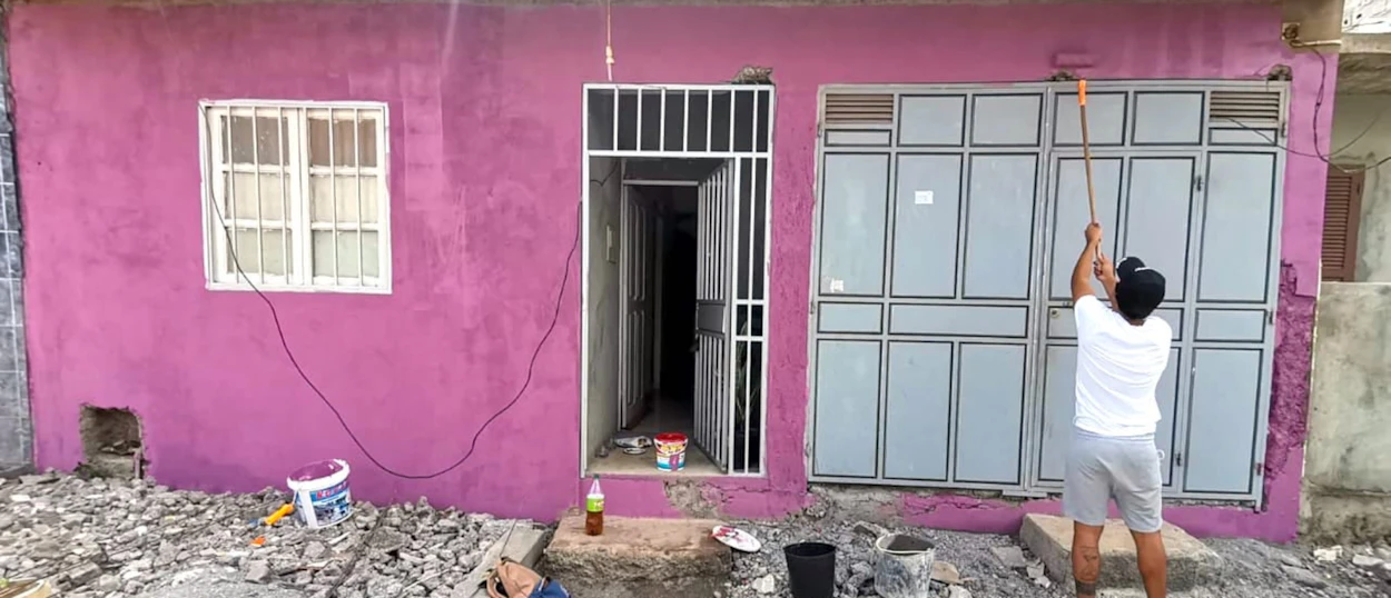 A person paints the exterior of a bright pink building with a long-handled roller, surrounded by debris.