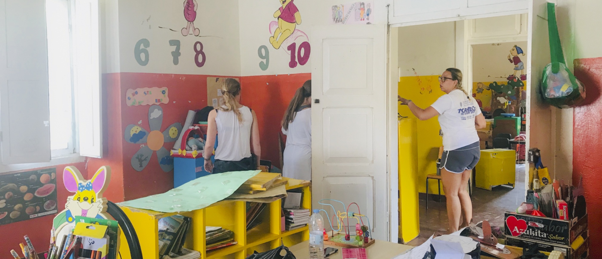 Volunteers paint yellow furniture and walls in a bright classroom with children's decorations.