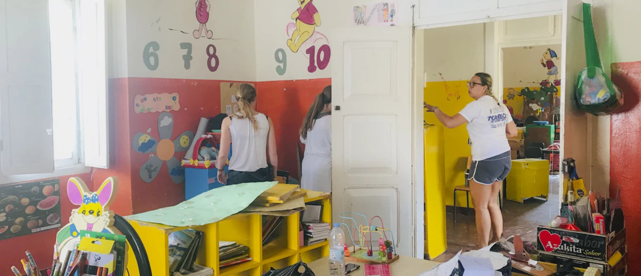 Volunteers paint yellow furniture and walls in a bright classroom with children's decorations.