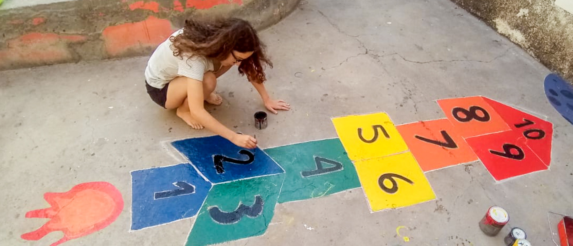 A young woman kneels, painting numbers onto a vibrant rocket-shaped hopscotch game on concrete ground.