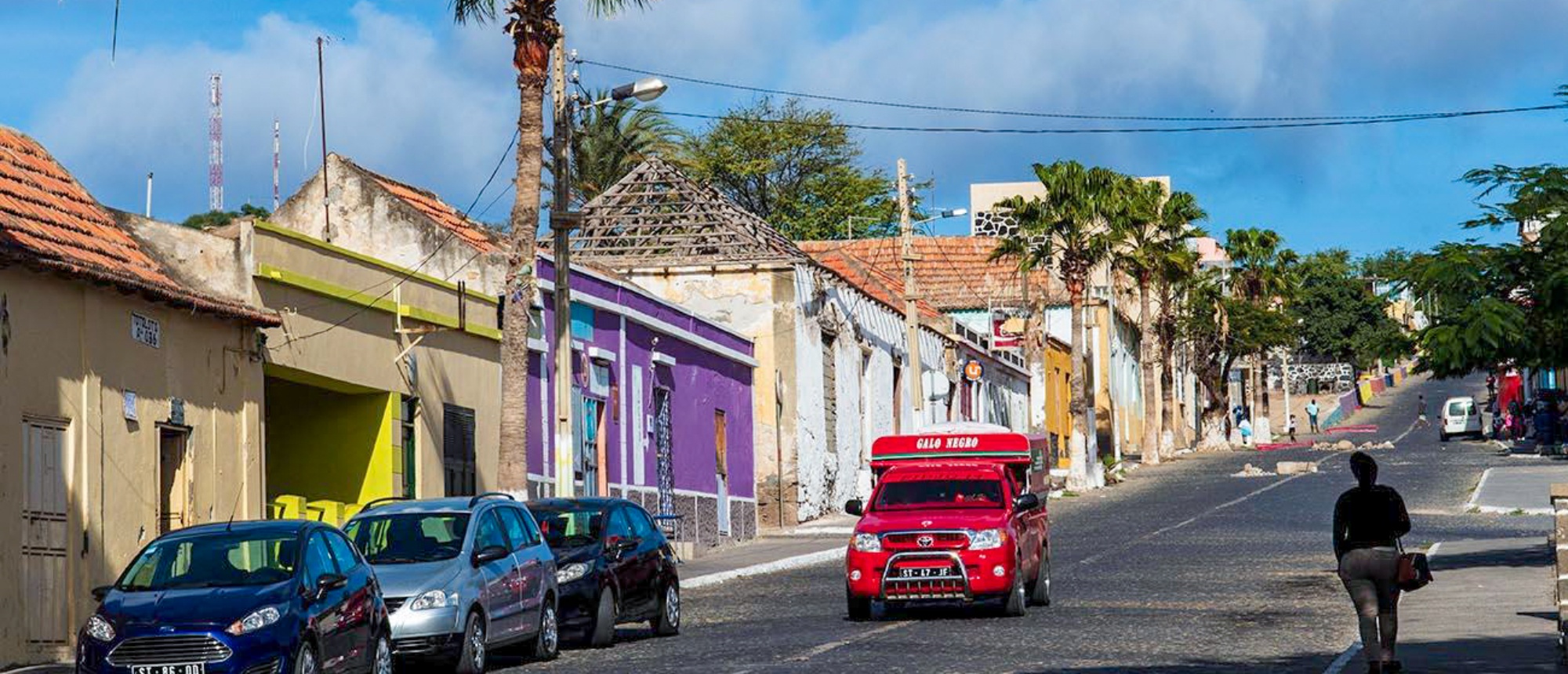 A vibrant street scene in Cape Verde with colorful buildings, parked cars, and a red taxi.