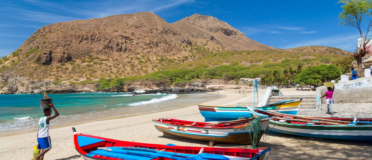 Colorful fishing boats on a sunny beach with people carrying baskets, backed by arid mountains.