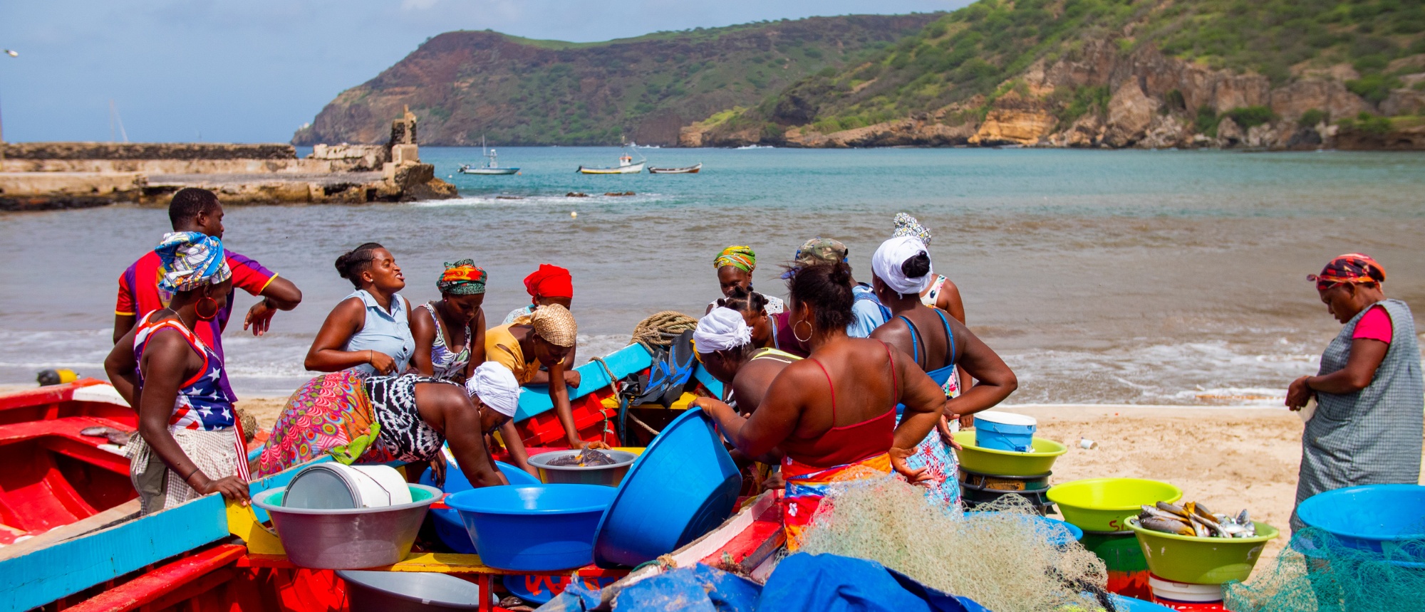 Women and men sort fresh fish and fishing nets on a busy beach by a calm bay.