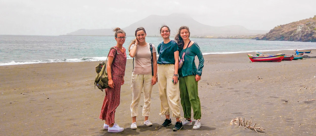 Four smiling women pose on a dark sand beach with mountains and fishing boats.