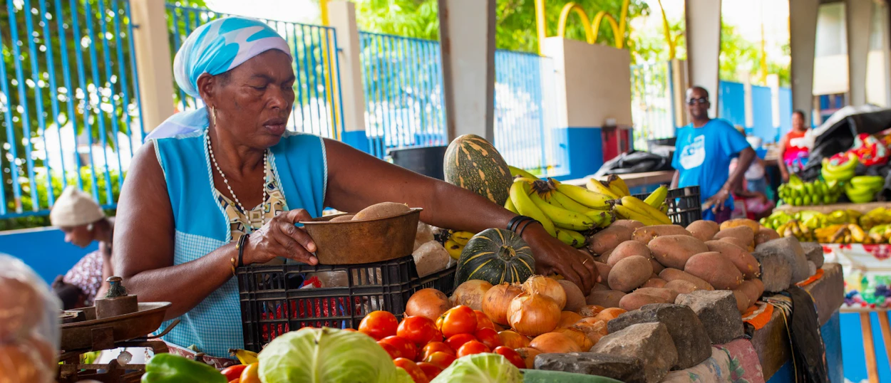 An older woman in a blue headscarf arranges fresh produce at a bustling market in Cape Verde.