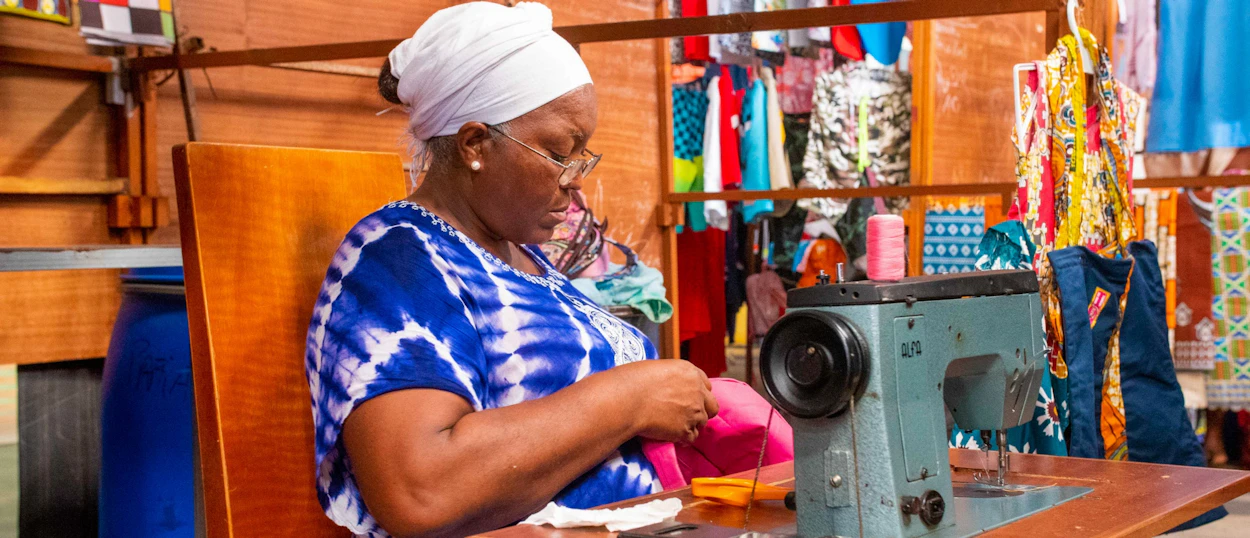A woman in a white headwrap sews colorful fabric at a machine in a Cape Verde market.