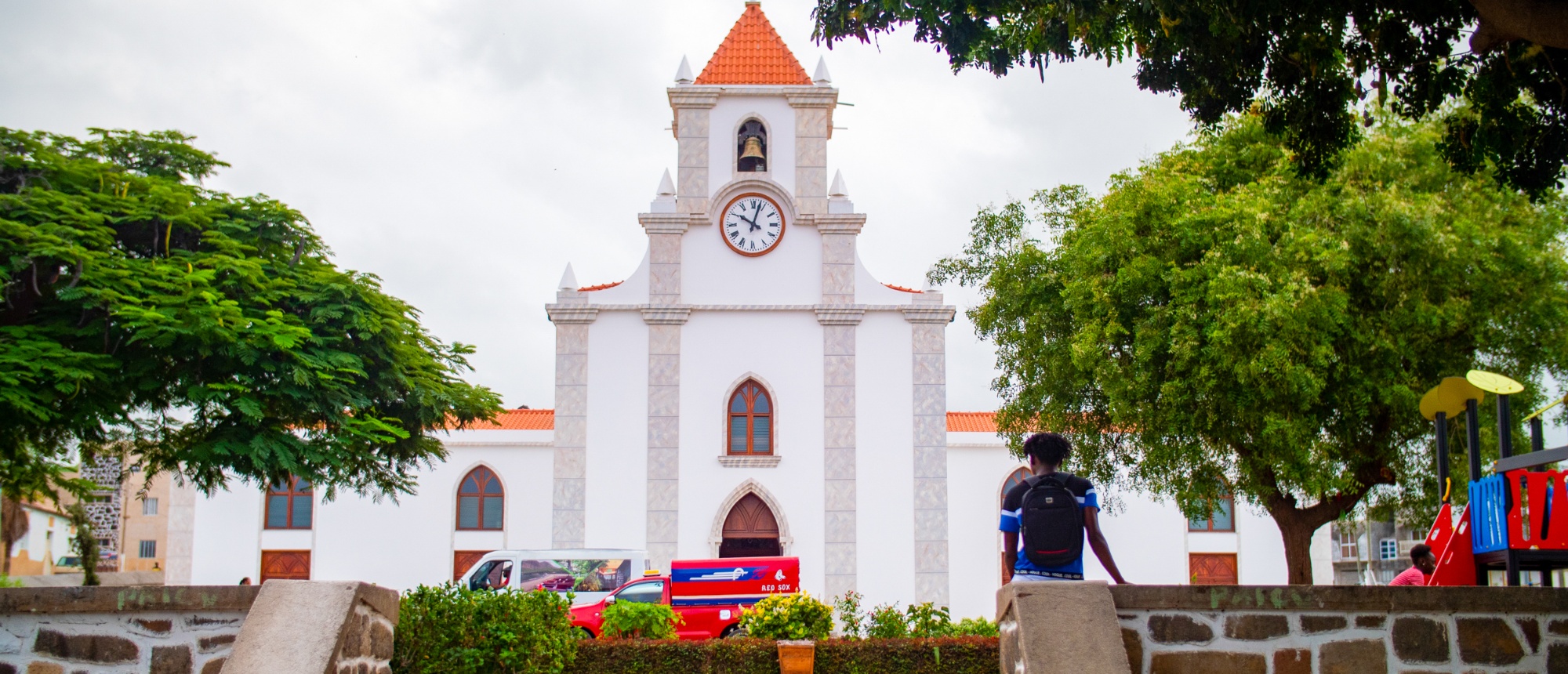 A person sits on a stone wall, looking at a white church with an orange roof in Terrafal.