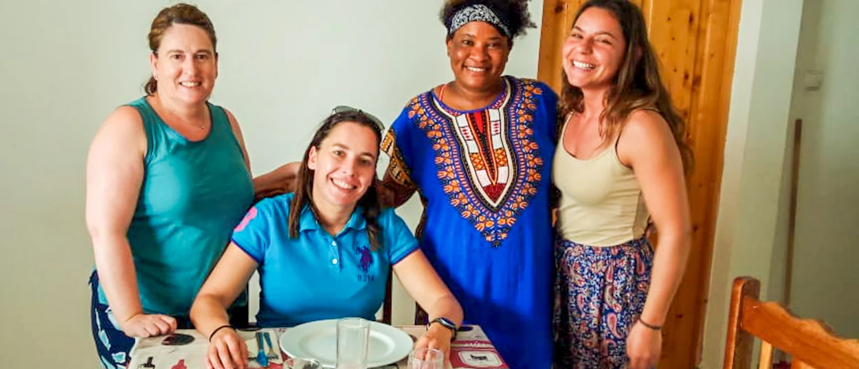 Four smiling women, one seated at a table, three standing, enjoying a friendly gathering indoors.