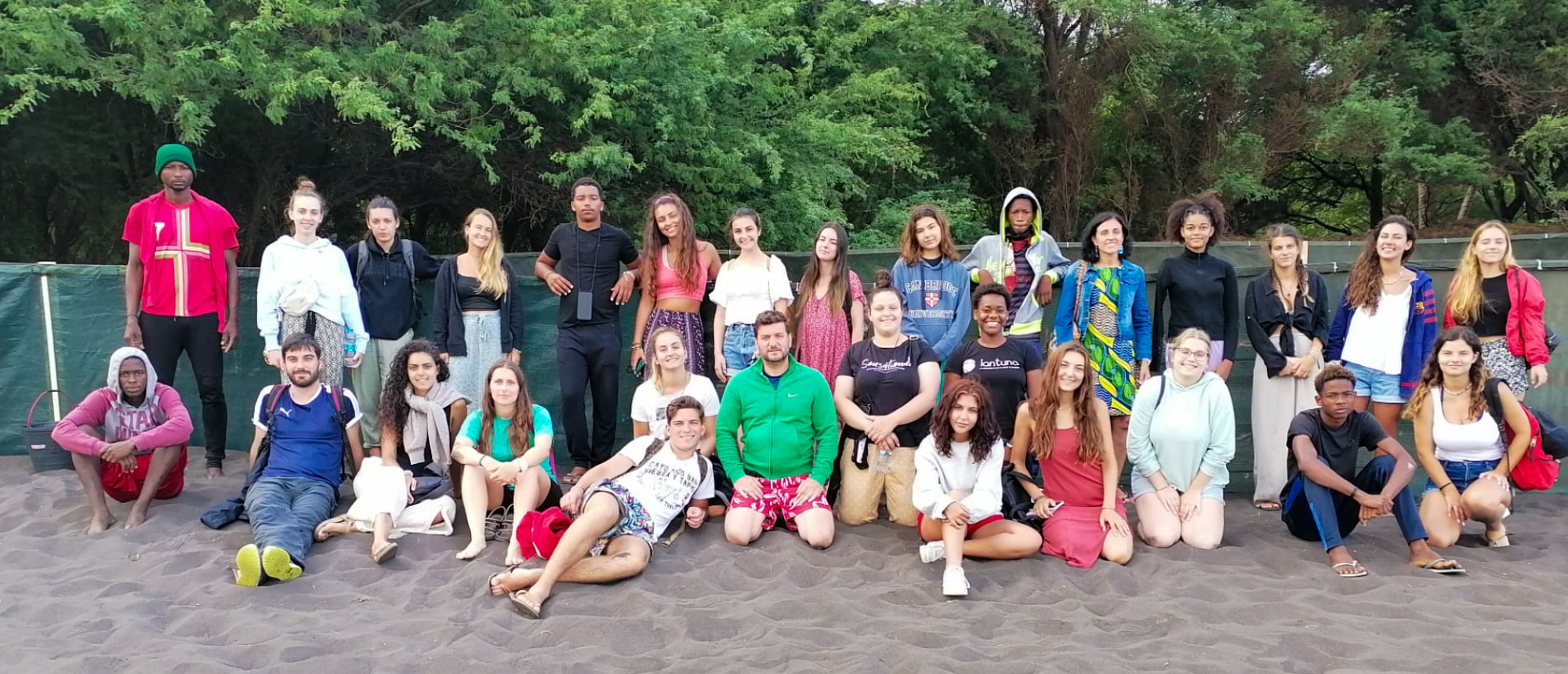 A diverse group of travellers smiling for a group photo on dark sand with green trees.
