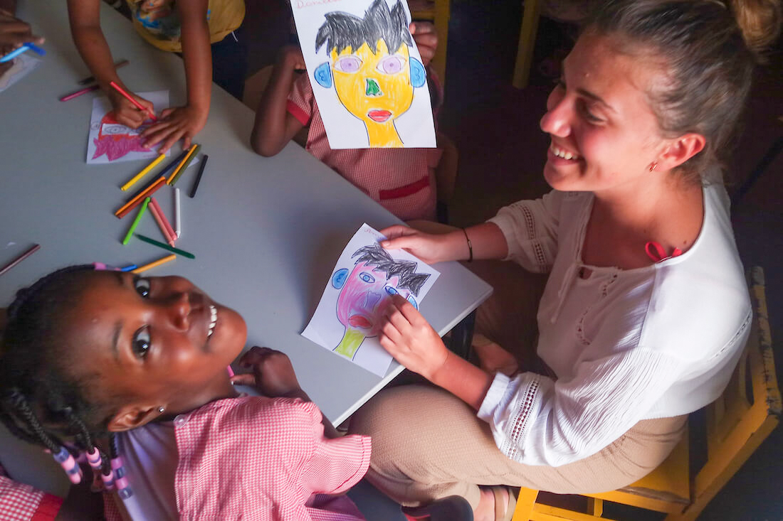 A smiling volunteer helps children draw colorful portraits, one child looks up happily.