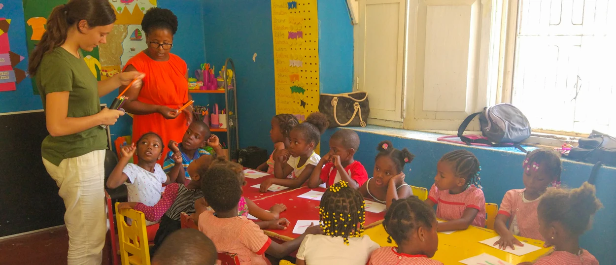 Two women teaching a group of young children seated at colorful tables in a bright classroom.