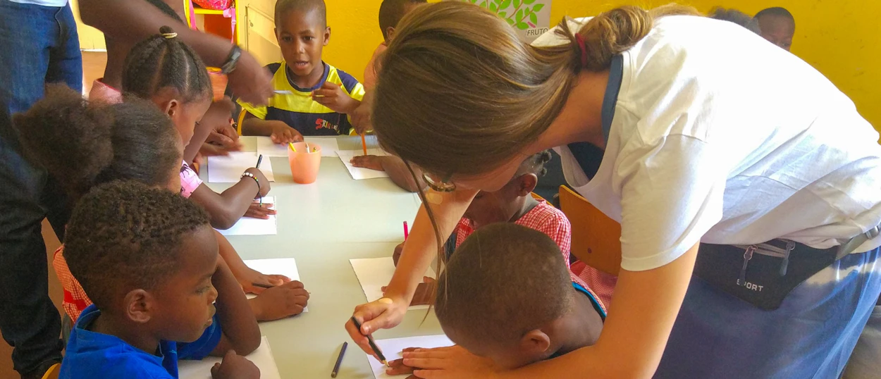 A volunteer helps a group of young children draw and color at a table in a bright classroom.