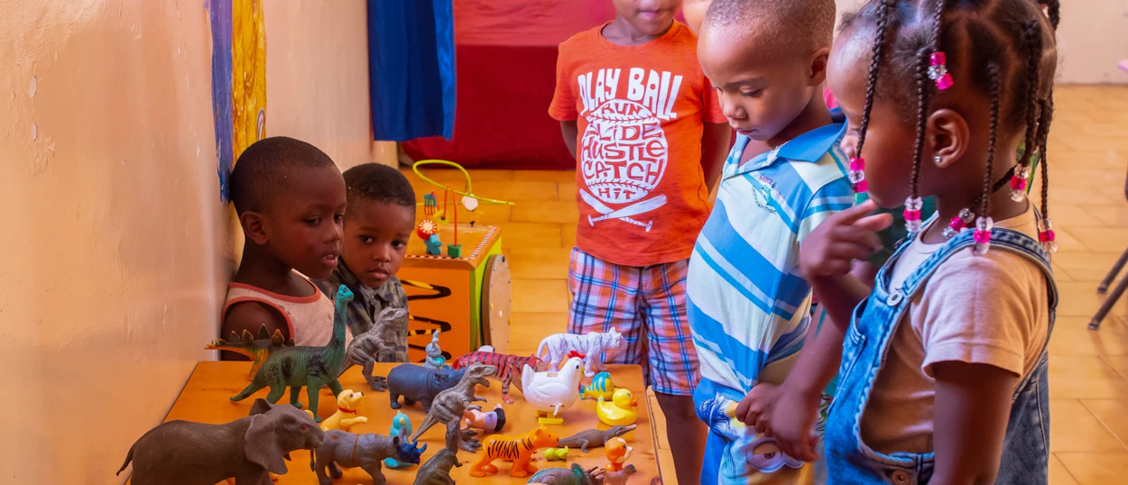 Curious kindergarten children gather around a table, exploring a variety of animal and dinosaur toys.