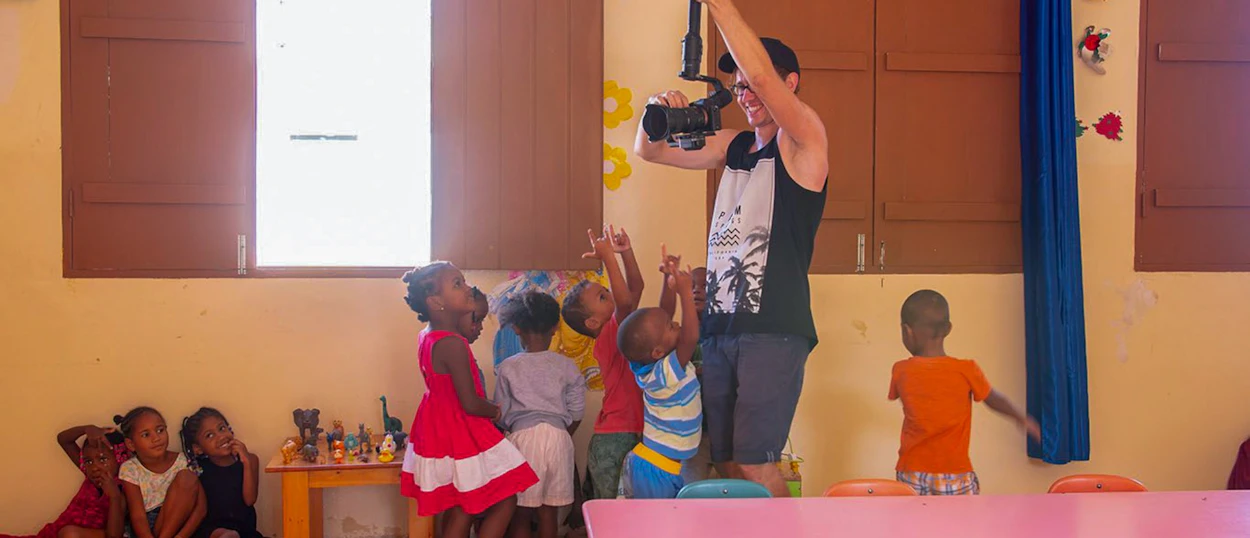 An adult films a group of excited children reaching up and playing in a cheerful classroom.