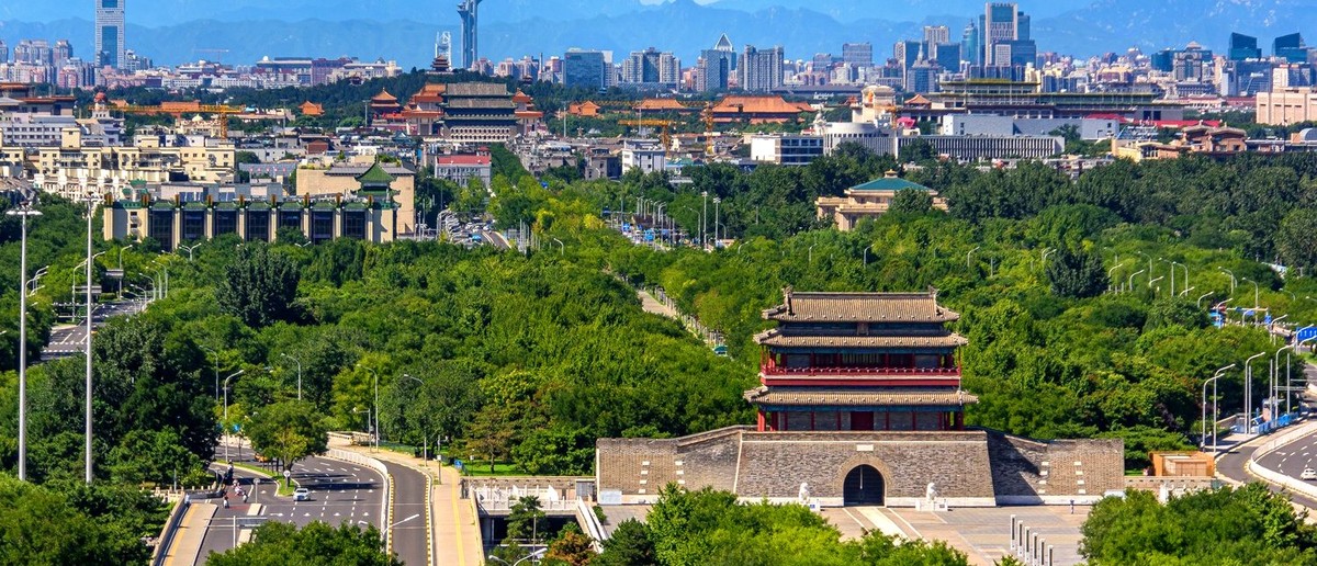 A traditional city gate tower stands in green parkland, with Beijing's skyline and mountains beyond.