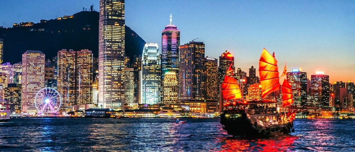 A red-sailed junk boat crosses Victoria Harbour beneath Hong Kong's lit-up skyscrapers at dusk.