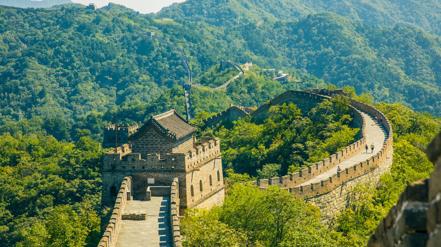 Tourists walk along the ancient Great Wall of China, winding through lush green mountains.