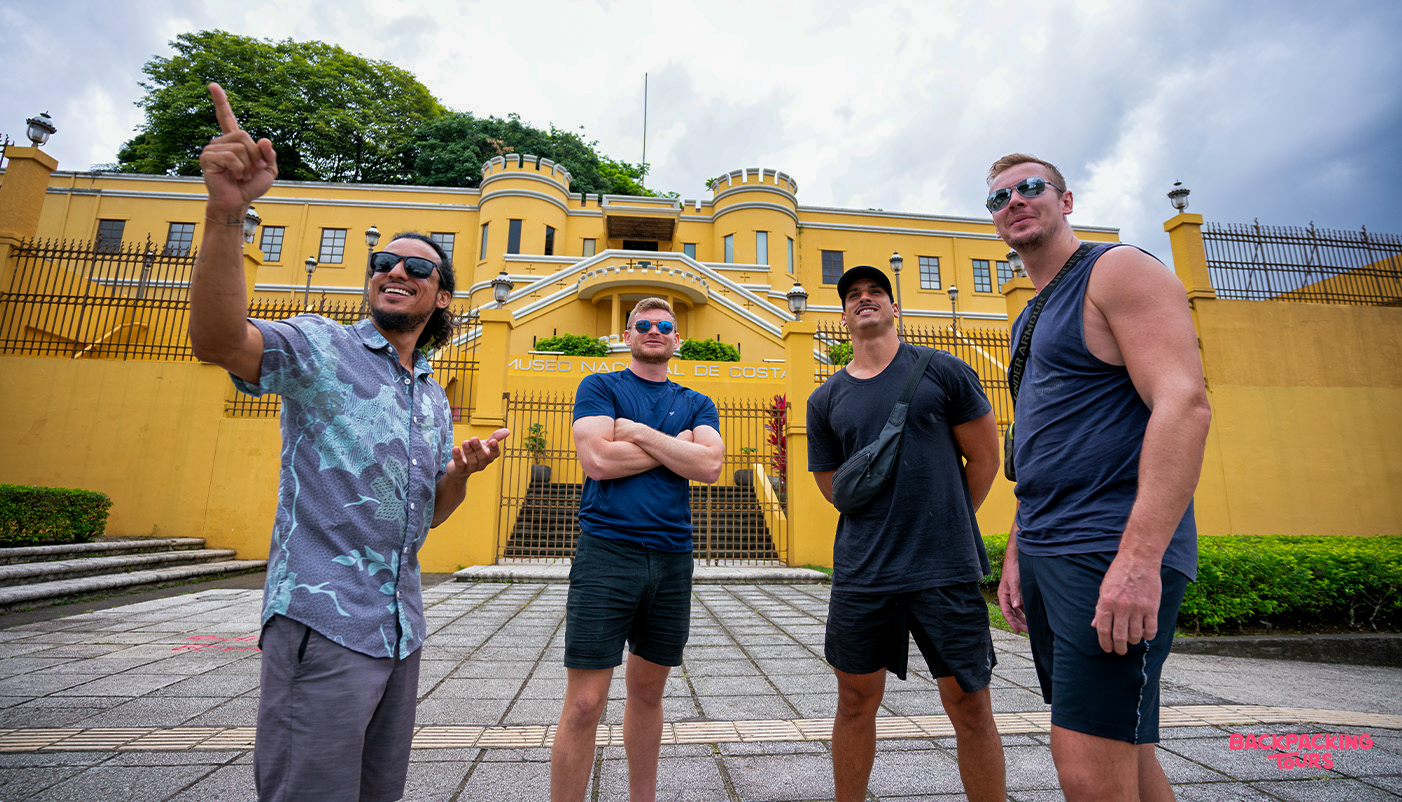 Four men on a guided city tour in San Jos es, pointing at a vibrant yellow historic building.