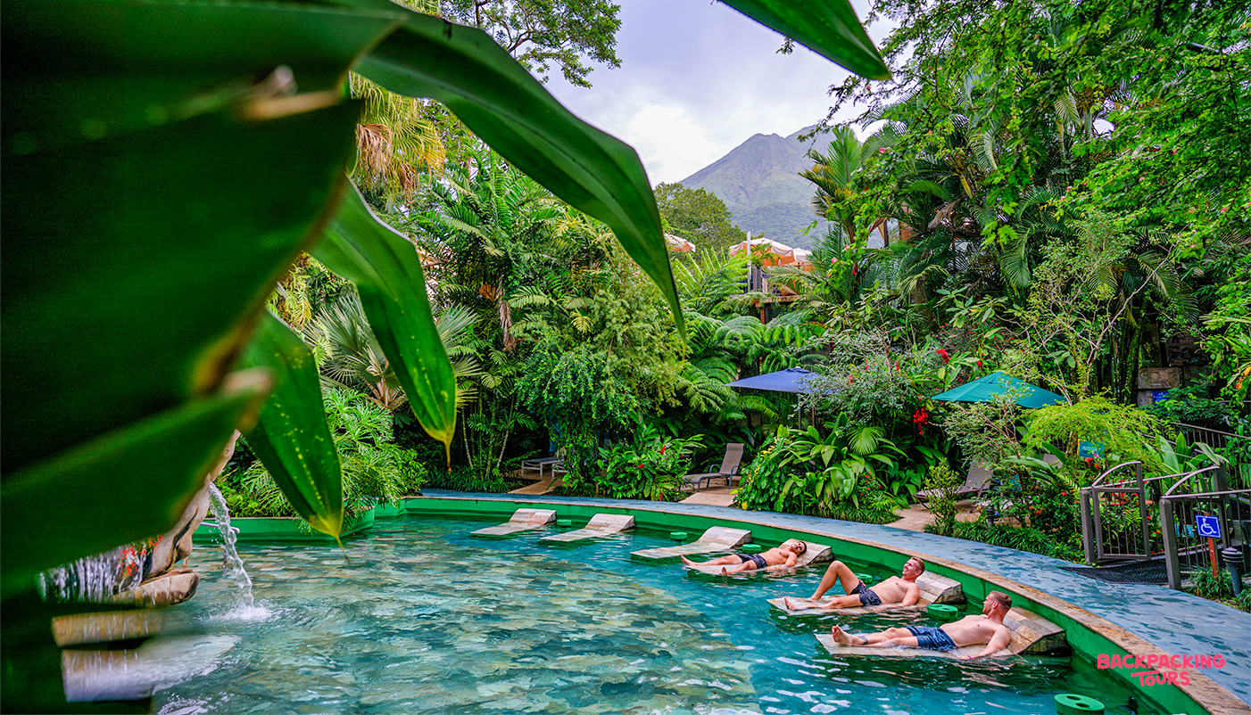 Travelers relax in natural hot springs surrounded by lush Costa Rican rainforest with a distant volcano.