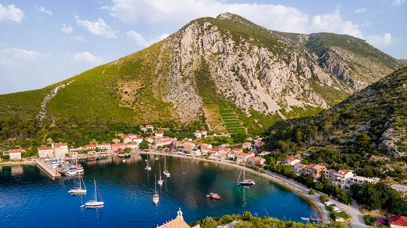 Trstenik, Croatia, a charming coastal village with sailboats in a blue bay, backed by a green mountain.
