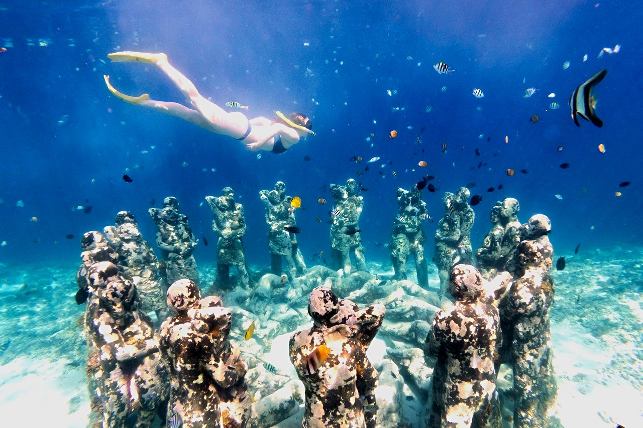 A snorkeller glides above rows of underwater statues off the Gili Islands, surrounded by tropical fish.
