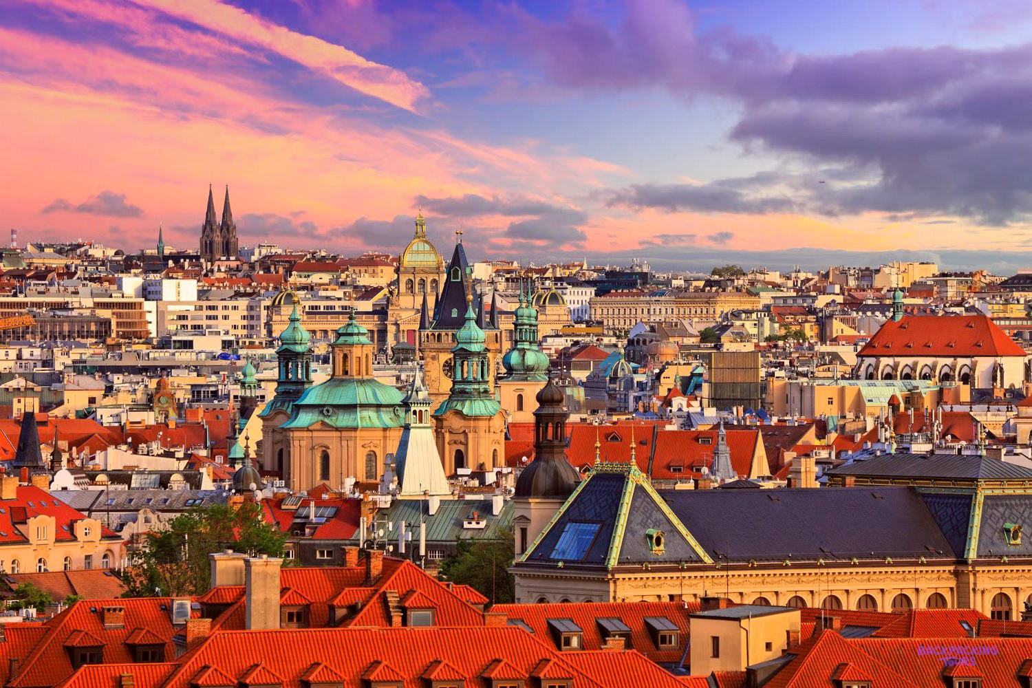 Red rooftops, green domes and church spires stretch across Prague under a pink sunset sky.