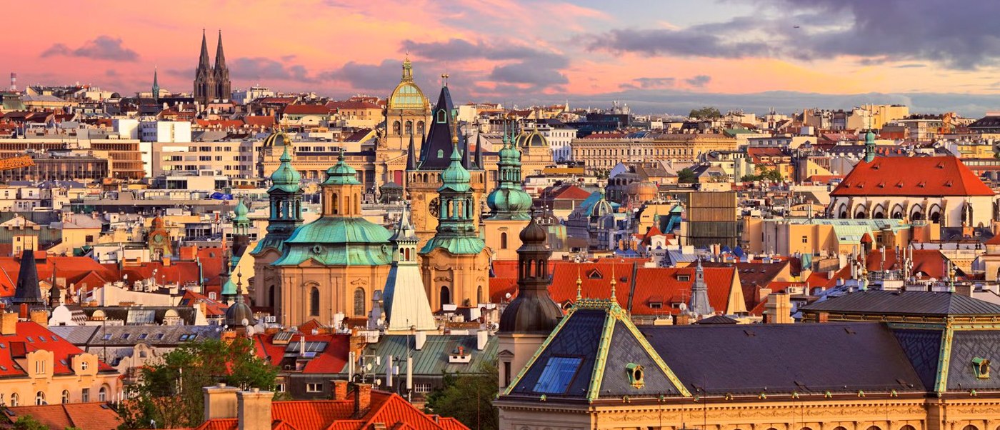 Red rooftops, green domes and church spires stretch across Prague under a pink sunset sky.