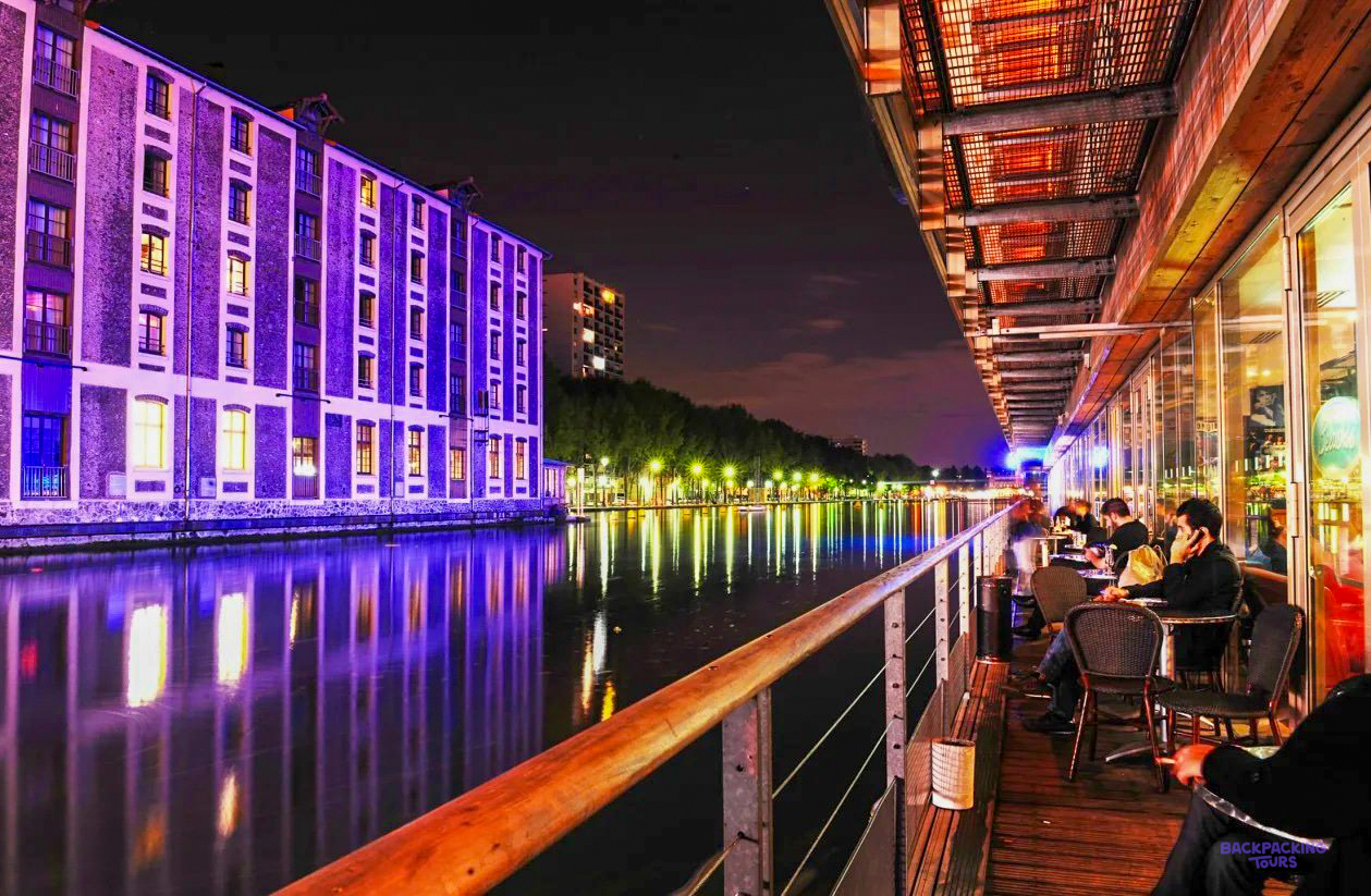 The terrace of the Paris hostel beside the canal at night, with the building opposite lit up in purple