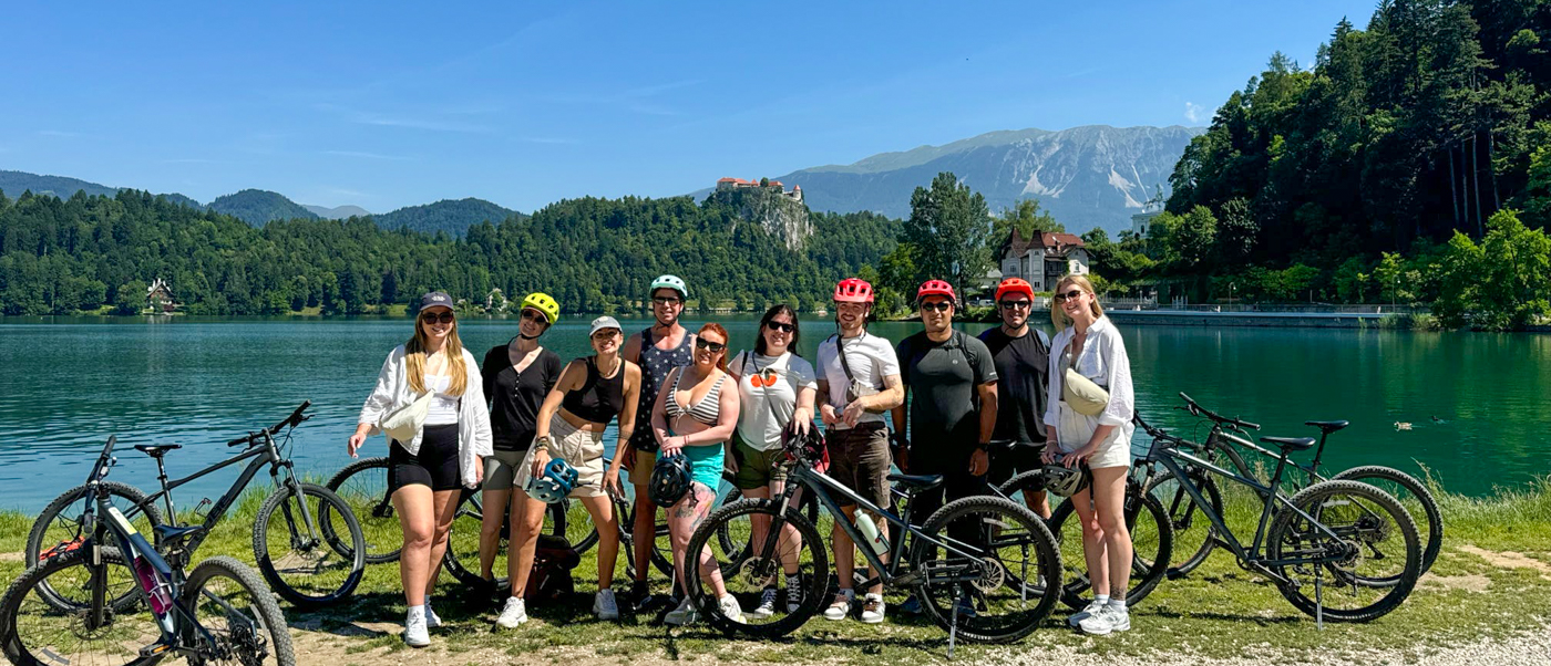 A group of cyclists stands with their bikes on the shore of Lake Bled.