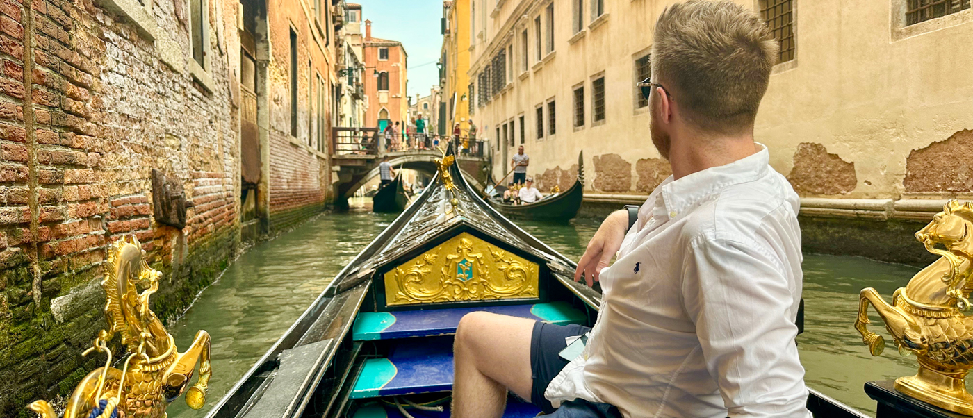 A traveller sits in a gold-trimmed gondola gliding down a narrow Venice canal.