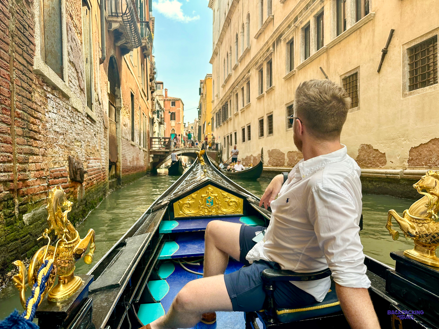 A traveller sits in a gold-trimmed gondola gliding down a narrow Venice canal.