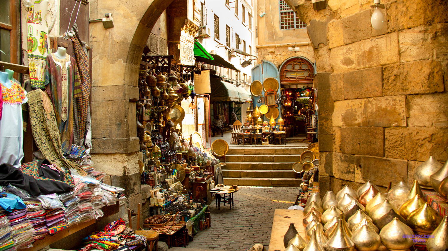 A bustling market alley in Cairo, with stalls overflowing with colourful textiles and shiny brass and copper goods.