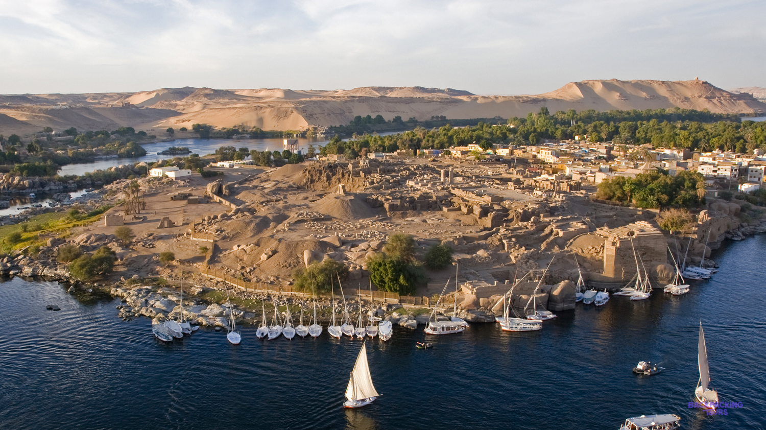 Boats sail around Elephantine Island in Aswan, Egypt, as the sun sets over the desert hills.