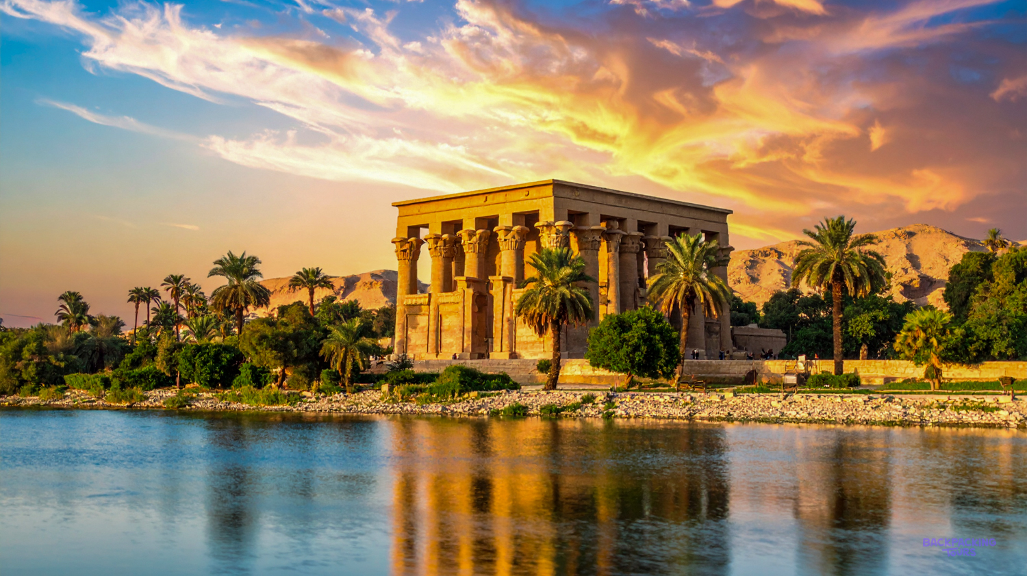 Ancient Philae Temple on the Nile River at sunset, surrounded by palm trees in Aswan.