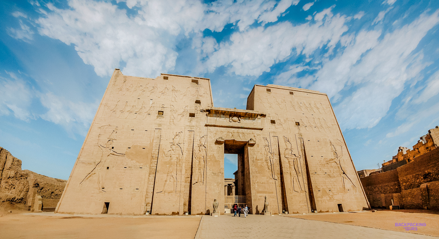 Visitors stand before the towering, carved entrance of the ancient Edfu Temple in Egypt under a blue sky.