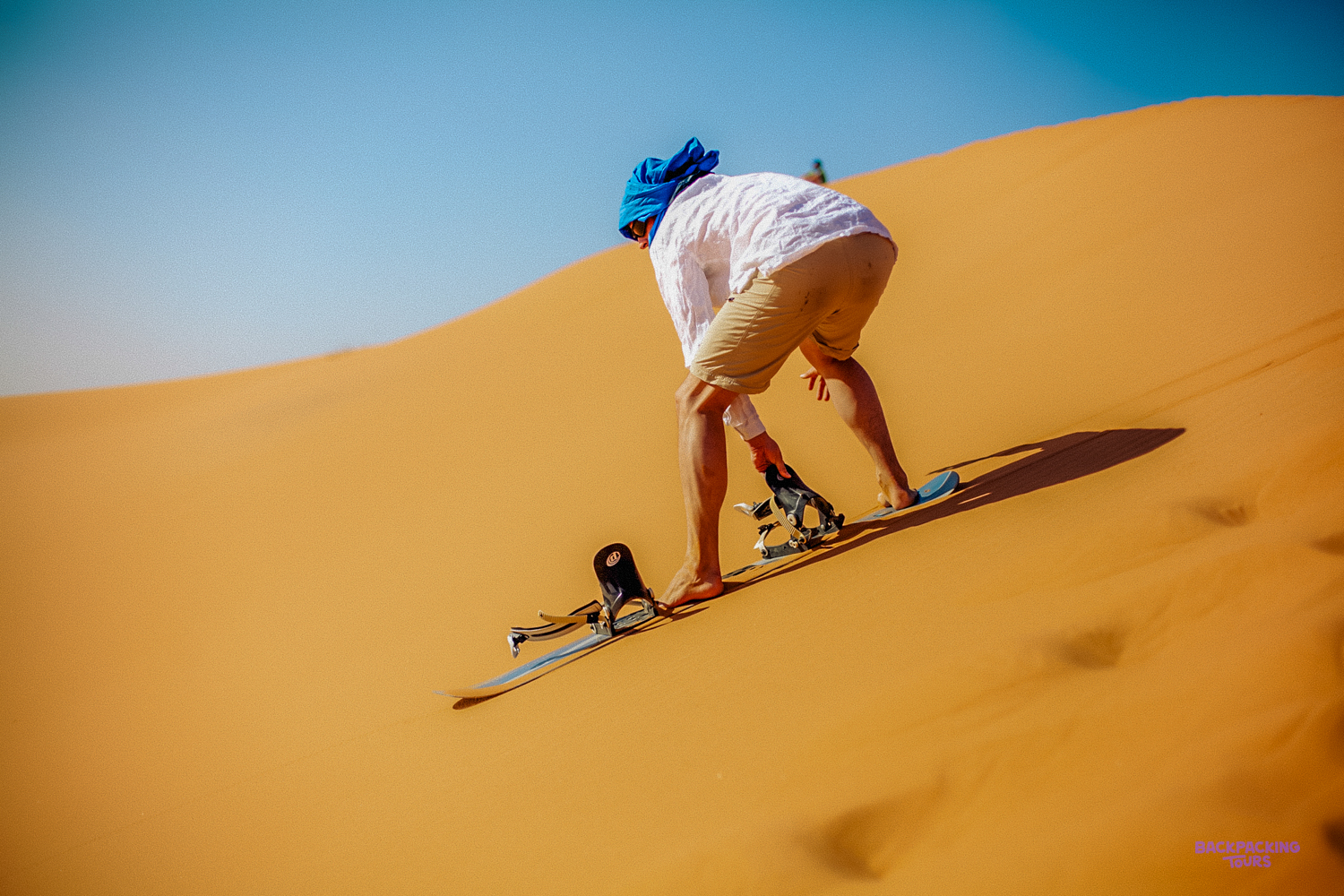 An adventurer sandboarding down a vast golden dune under a clear blue sky in Wadi al Hitan.
