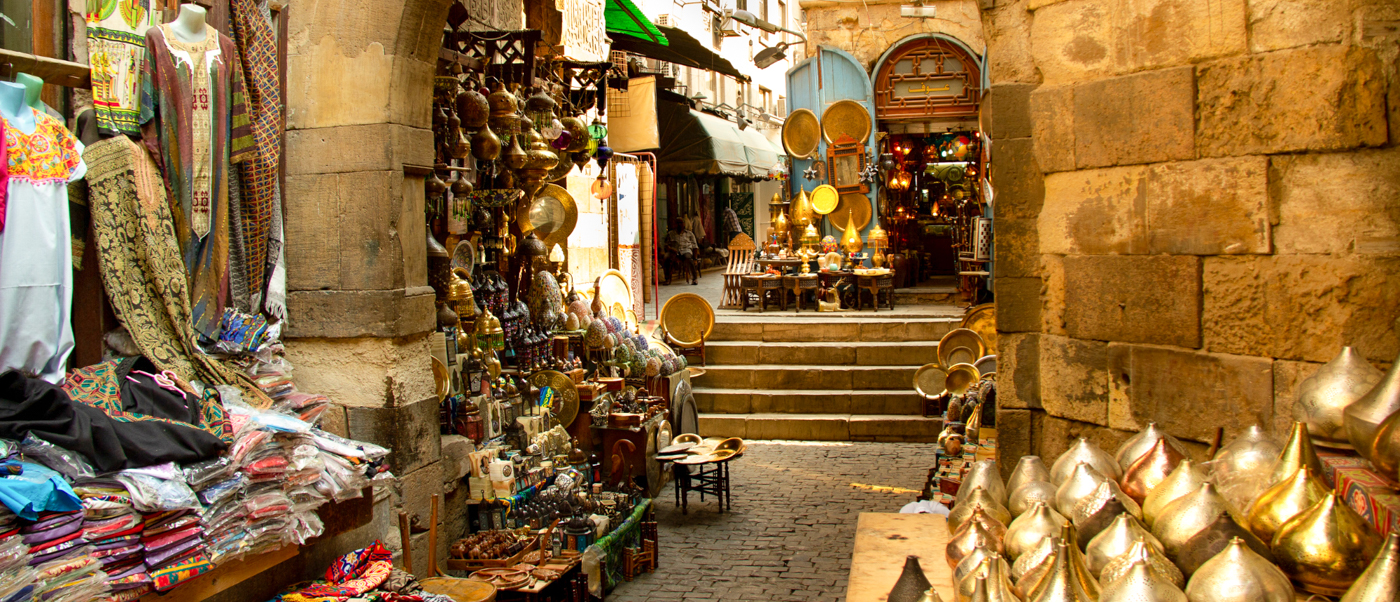 A vibrant market alley in Cairo, Egypt, filled with stalls selling colorful textiles and shining metalware.