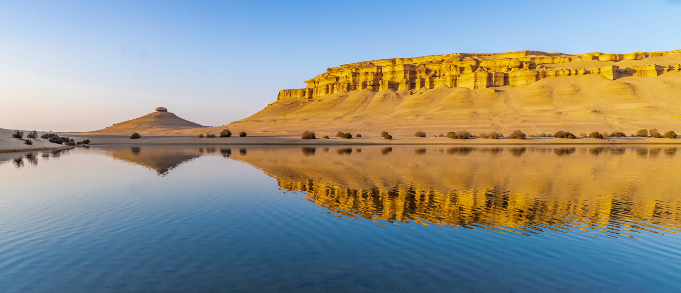 A serene desert lake reflects golden layered cliffs under a clear blue sky in Faiyum, Egypt.